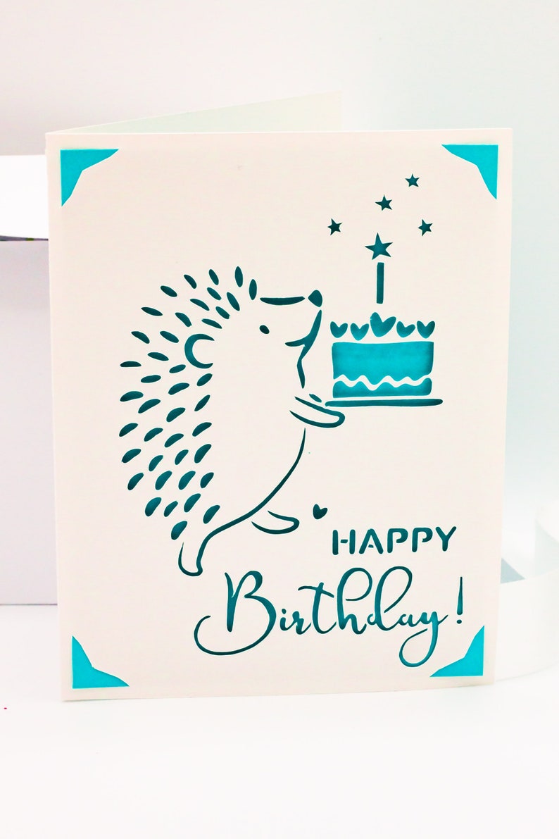 May include: A white card with a teal blue hedgehog holding a teal blue cake with a single candle and stars. The card says "Happy Birthday!" in teal blue lettering.