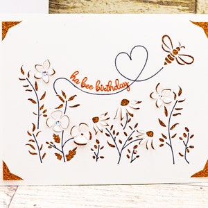 May include: A white card with a bee and flowers cut out. The card has the words "ha bee birthday" written in copper glitter.