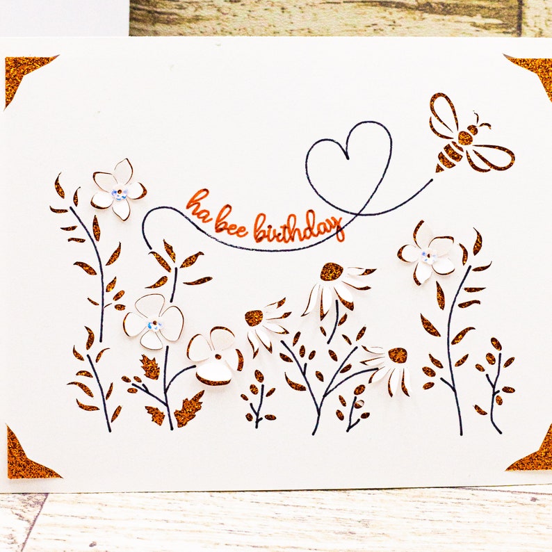 May include: A white card with a bee and flowers cut out. The card has the words "ha bee birthday" written in copper glitter.