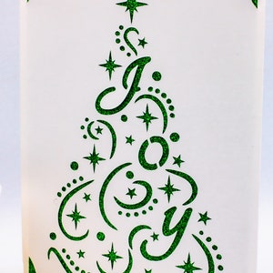 May include: A white card with a green glitter design of a Christmas tree with the word "Joy" inside. The tree is decorated with stars and crescent moons.