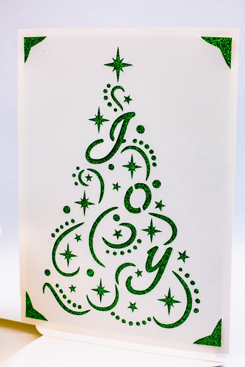 May include: A white card with a green glitter design of a Christmas tree with the word "Joy" inside. The tree is decorated with stars and crescent moons.