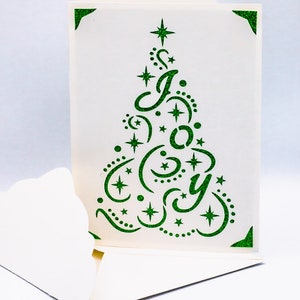 May include: A white card with a green glitter cut-out design of a Christmas tree with the word "JOY" inside. The card is folded in half and the inside is blank.