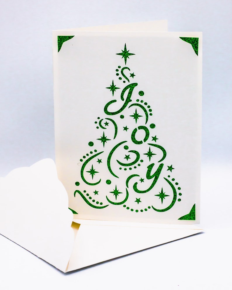 May include: A white card with a green glitter cut-out design of a Christmas tree with the word "JOY" inside. The card is folded in half and the inside is blank.