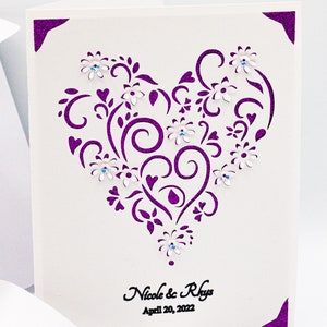 May include: A white card with a purple glitter border and a cut-out heart design. The heart is decorated with white flowers and rhinestones. The text "Nicole & Rhys April 20, 2022" is printed in black at the bottom of the card.