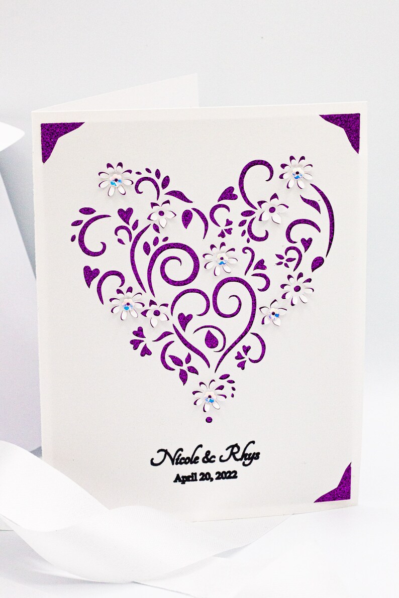 May include: A white card with a purple glitter border and a cut-out heart design. The heart is decorated with white flowers and rhinestones. The text "Nicole & Rhys April 20, 2022" is printed in black at the bottom of the card.
