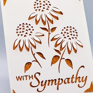 Puede incluir: Una tarjeta blanca con un borde de purpurina dorada y un dise&ntilde;o recortado de tres flores blancas con tallos y hojas marrones. El texto "With Sympathy" est&aacute; escrito en purpurina dorada en la tarjeta.
