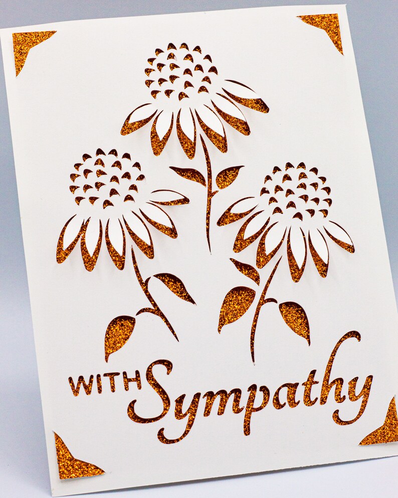 Puede incluir: Una tarjeta blanca con un borde de purpurina dorada y un dise&ntilde;o recortado de tres flores blancas con tallos y hojas marrones. El texto "With Sympathy" est&aacute; escrito en purpurina dorada en la tarjeta.