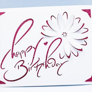 May include: A white card with a pink glitter design that says "Happy Birthday" with a white paper flower in the center.