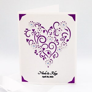 May include: A white card with a purple heart-shaped design featuring intricate swirls and flowers. The heart is decorated with small, clear rhinestones. The text "Nicole & Rhys" and "April 20, 2022" is printed below the heart.