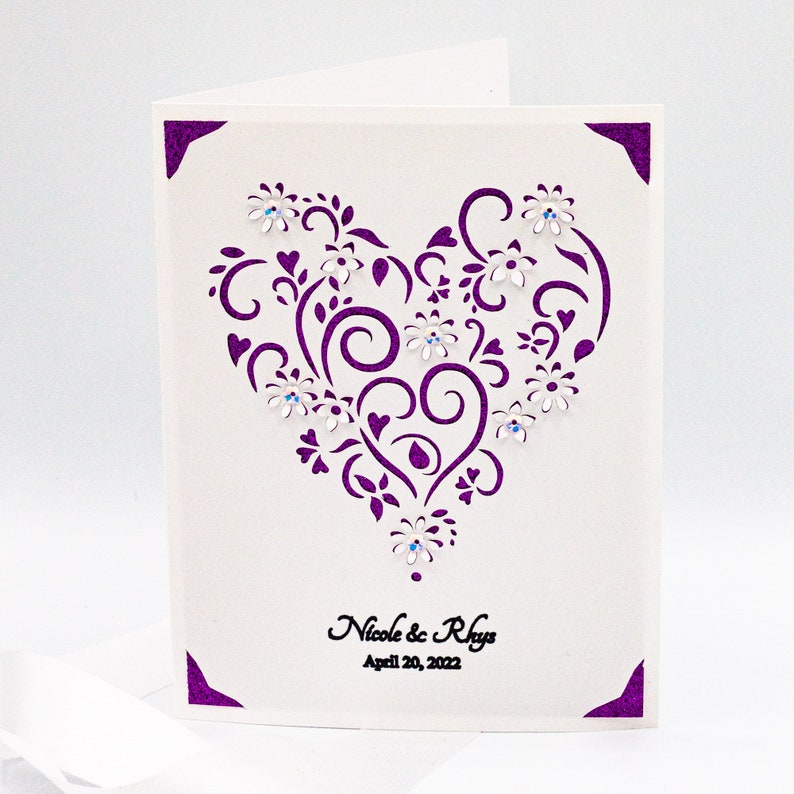 May include: A white card with a purple heart-shaped design featuring intricate swirls and flowers. The heart is decorated with small, clear rhinestones. The text "Nicole & Rhys" and "April 20, 2022" is printed below the heart.