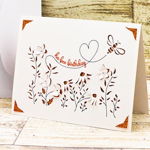 May include: A white card with a brown border and a cut-out design of flowers, leaves, and a bee. The text "ha bee birthday" is written in orange cursive.