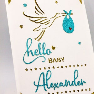 May include: A white card with gold glitter corners and a blue glitter stork carrying a blue glitter baby bundle. The card says "hello baby Alexander" in blue glitter.