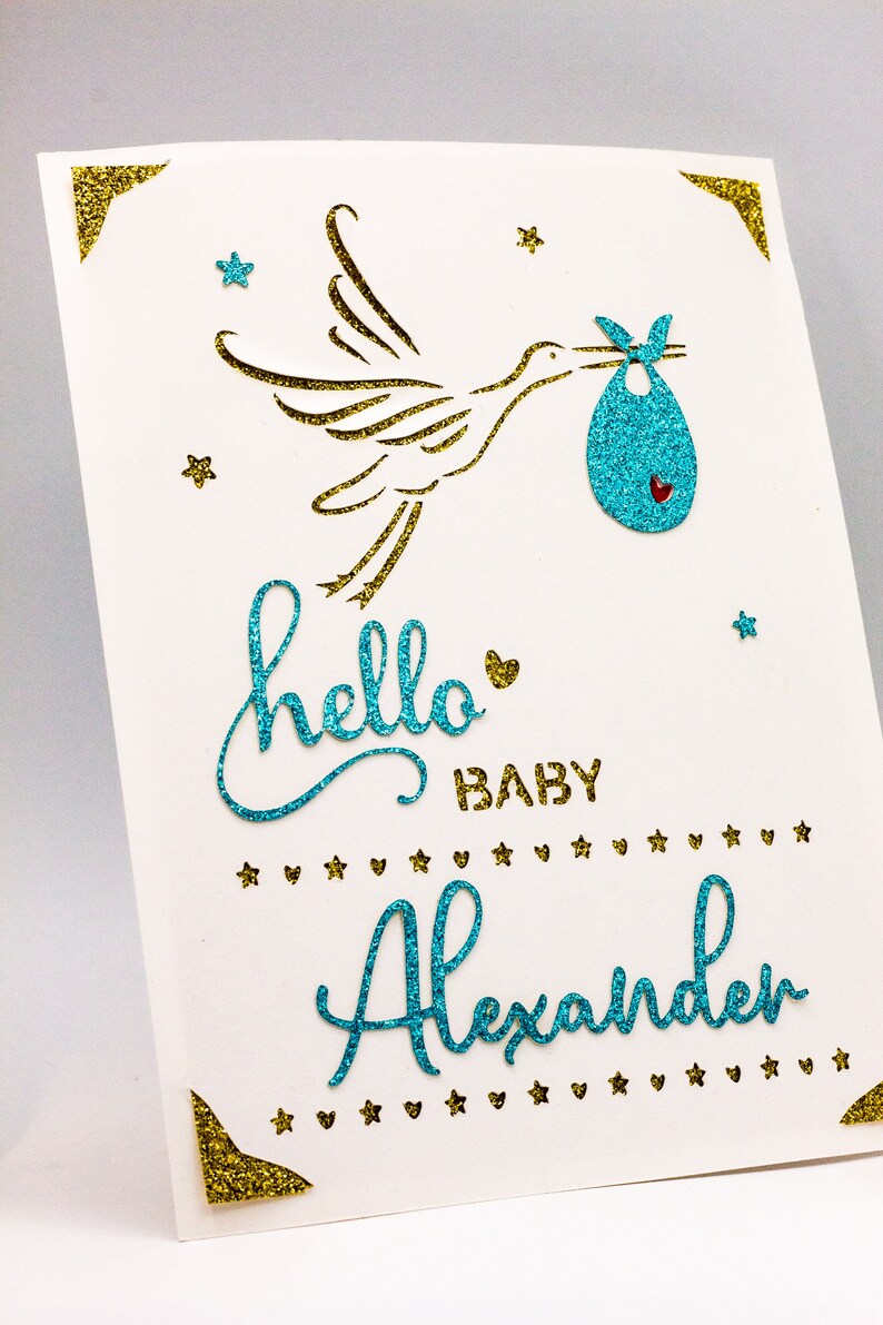 May include: A white card with gold glitter corners and a blue glitter stork carrying a blue glitter baby bundle. The card says "hello baby Alexander" in blue glitter.