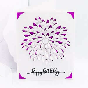 May include: A white card with a pink glitter border and a white paper flower in the center. The flower is made of many layers of paper that have been cut out to create a 3D effect. The card also has the words "happy birthday" written in black ink.