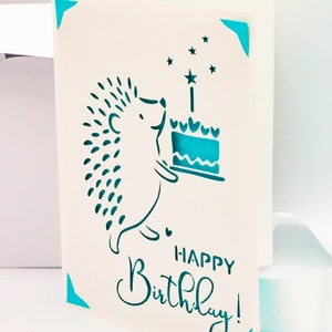 May include: A white card with a blue hedgehog holding a blue cake with a single candle and stars. The text "Happy Birthday!" is written in blue cursive.
