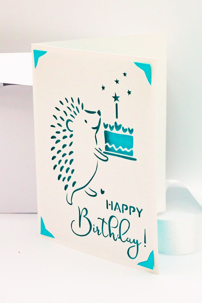 May include: A white card with a blue hedgehog holding a blue cake with a single candle and stars. The text "Happy Birthday!" is written in blue cursive.