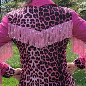Puede incluir: Chaqueta rosa y negra con estampado de leopardo con flecos y mangas rosas. La chaqueta tiene un estilo western cl&aacute;sico con cuello y canes&uacute; en forma de V en la espalda. Los flecos est&aacute;n adornados con peque&ntilde;os detalles brillantes.