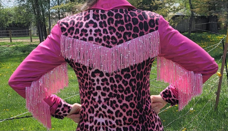 Puede incluir: Chaqueta rosa y negra con estampado de leopardo con flecos y mangas rosas. La chaqueta tiene un estilo western cl&aacute;sico con cuello y canes&uacute; en forma de V en la espalda. Los flecos est&aacute;n adornados con peque&ntilde;os detalles brillantes.