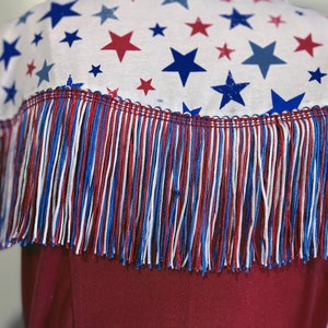 May include: A white fabric with a pattern of red and blue stars, trimmed with red, white, and blue fringe. The fringe is attached to a burgundy garment, creating a patriotic design.