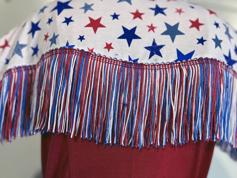 May include: A white fabric with a pattern of red and blue stars, trimmed with red, white, and blue fringe. The fringe is attached to a burgundy garment, creating a patriotic design.