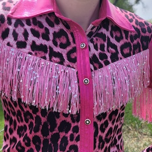 Puede incluir: Camisa rosa y negra con estampado de leopardo, flecos y lentejuelas. La camisa tiene cuello y mangas largas. Los flecos est&aacute;n en el pecho y las mangas. La camisa est&aacute; abotonada por delante.