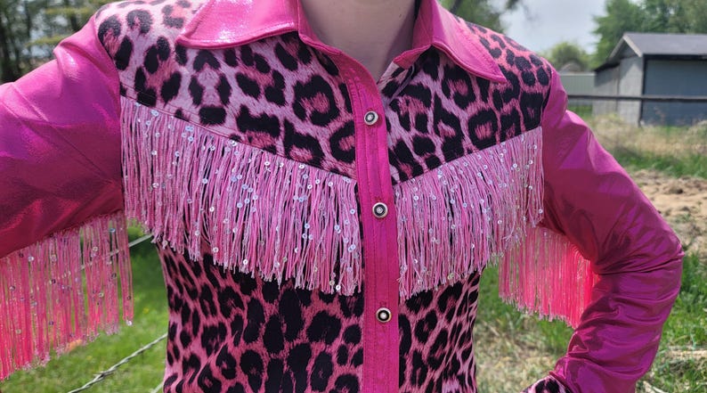 Puede incluir: Camisa rosa y negra con estampado de leopardo, flecos y lentejuelas. La camisa tiene cuello y mangas largas. Los flecos est&aacute;n en el pecho y las mangas. La camisa est&aacute; abotonada por delante.