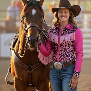 Puede incluir: Una mujer con sombrero de vaquero marr&oacute;n y camisa rosa con estampado de leopardo y flecos, junto a un caballo marr&oacute;n. Lleva vaqueros azules y una hebilla plateada. El caballo tiene una marca blanca en la frente.