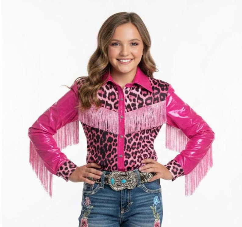 Puede incluir: Una camisa con estampado de leopardo rosa y negro con flecos y mangas largas rosas. La camisa se combina con vaqueros azules y un cintur&oacute;n decorativo con detalles turquesa. El conjunto es de estilo western.