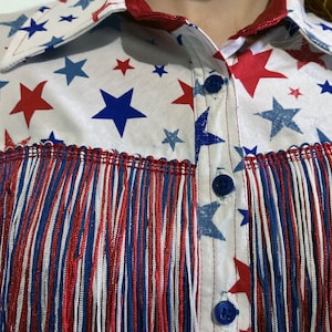 May include: A white shirt with a pattern of red and blue stars. The shirt has a collar and blue buttons. The bottom of the shirt has red, white, and blue fringe.