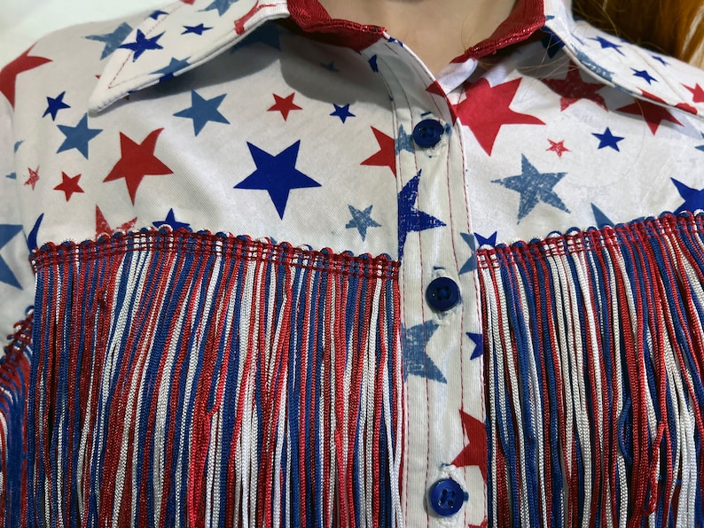 May include: A white shirt with a pattern of red and blue stars. The shirt has a collar and blue buttons. The bottom of the shirt has red, white, and blue fringe.