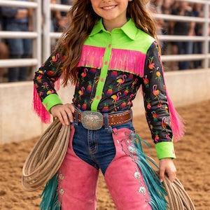 Op de afbeelding: Een persoon draagt een strooien cowboyhoed en een zwart shirt met een flamingo- en ananaspatroon. Het shirt heeft een neon groene kraag en manchetten en roze franjes. Ze dragen een spijkerbroek, roze chaps met turquoise franjes en een bruine riem.
