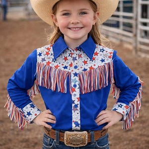 Op de afbeelding: Een jong meisje draagt een strooien cowboyhoed en een blauw shirt met rode, witte en blauwe sterren en franjes. Ze draagt ook een bruine riem met een decoratieve gesp en een blauwe spijkerbroek.