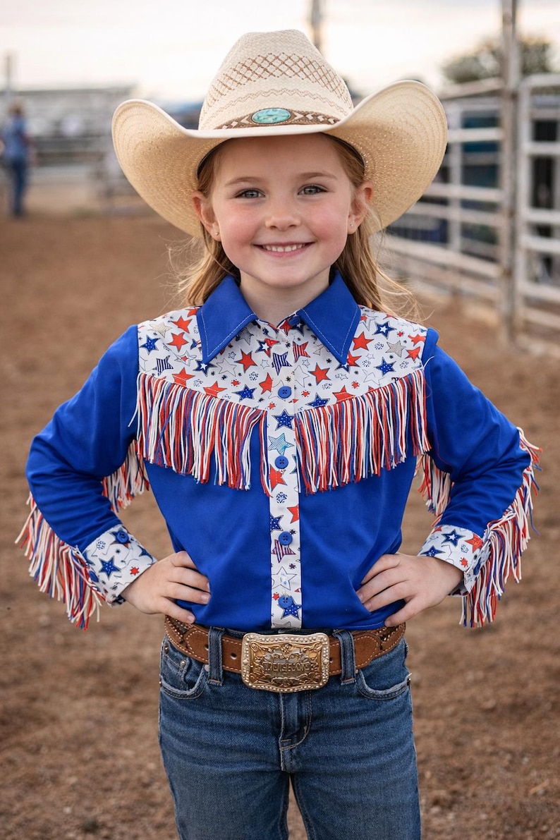 Op de afbeelding: Een jong meisje draagt een strooien cowboyhoed en een blauw shirt met rode, witte en blauwe sterren en franjes. Ze draagt ook een bruine riem met een decoratieve gesp en een blauwe spijkerbroek.