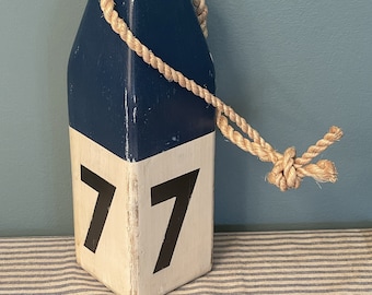 May include: A wooden nautical buoy with a blue top and white bottom. The buoy is numbered "77" and has a rope loop at the top.