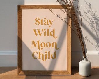 May include: A framed print with a light pink background and the text "Stay Wild Moon Child" in a gold color. The frame is made of wood and has a natural finish. A vase with dried flowers is in the foreground.