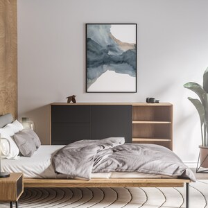 May include: A bedroom with a wooden bed frame, a grey duvet, and a bedside table with a lamp. There is a wooden cabinet with shelves and a black drawer. A large plant in a metal pot is in the corner of the room. A framed abstract watercolour painting hangs on the wall.