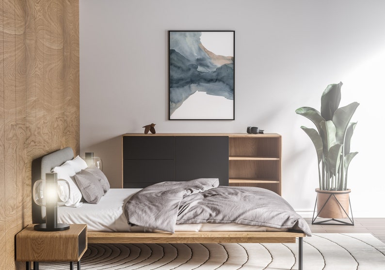 May include: A bedroom with a wooden bed frame, a grey duvet, and a bedside table with a lamp. There is a wooden cabinet with shelves and a black drawer. A large plant in a metal pot is in the corner of the room. A framed abstract watercolour painting hangs on the wall.