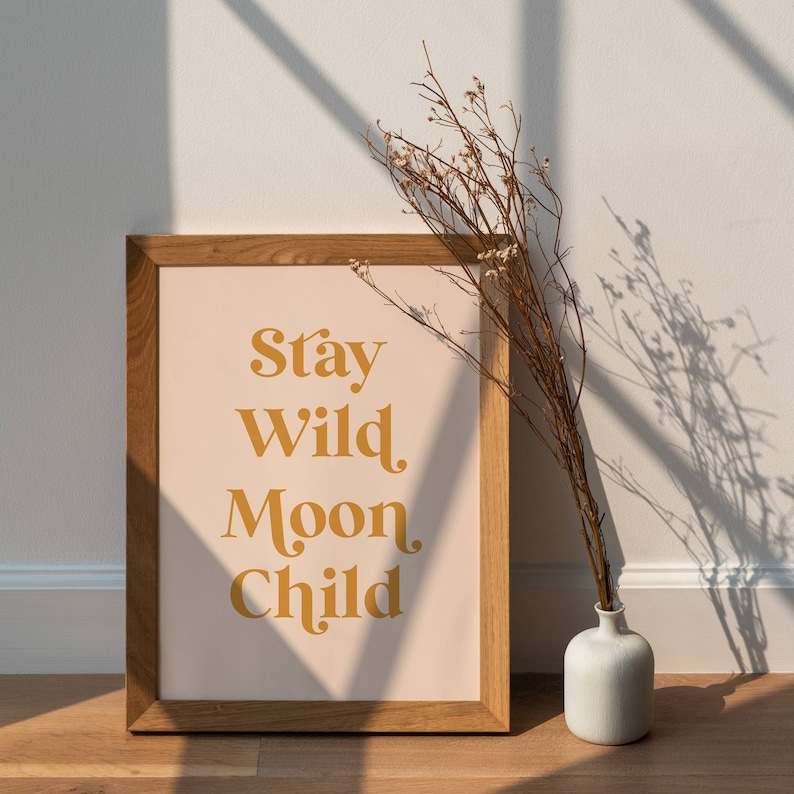 May include: A framed print with a light pink background and the text "Stay Wild Moon Child" in a gold color. The frame is made of wood and has a natural finish. A vase with dried flowers is in the foreground.