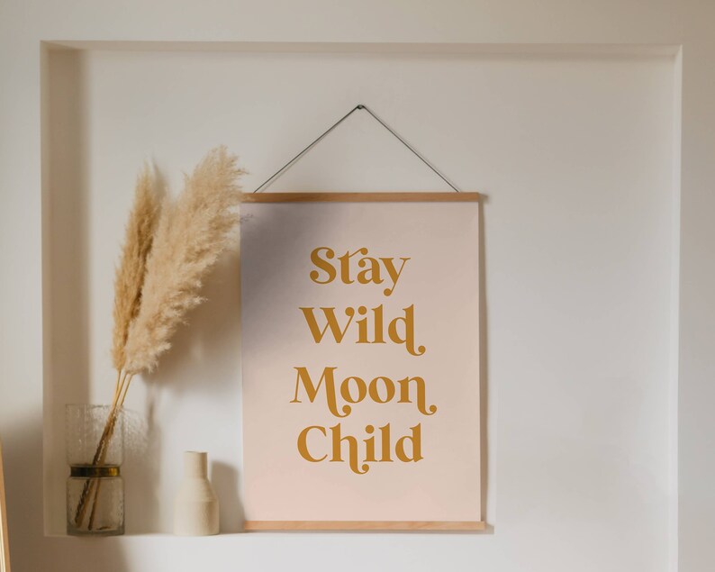 May include: A framed print with a pink background and gold text that reads "Stay Wild Moon Child". The print is hanging on a white wall with a dried pampas grass arrangement in a clear glass vase to the left.