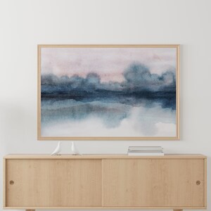 K&ouml;nnte beinhalten: Ein gerahmtes Aquarellbild einer abstrakten Landschaft in Blau und Rosa. Das Bild h&auml;ngt an einer wei&szlig;en Wand &uuml;ber einem Holzschrank.