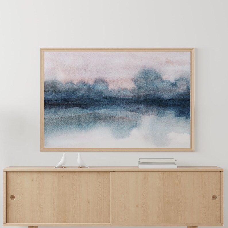 K&ouml;nnte beinhalten: Ein gerahmtes Aquarellbild einer abstrakten Landschaft in Blau und Rosa. Das Bild h&auml;ngt an einer wei&szlig;en Wand &uuml;ber einem Holzschrank.