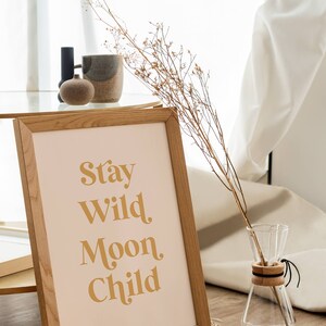 May include: A framed print with the text "Stay Wild Moon Child" in a gold font on a light pink background. The frame is made of light brown wood and is sitting on a woven rug.