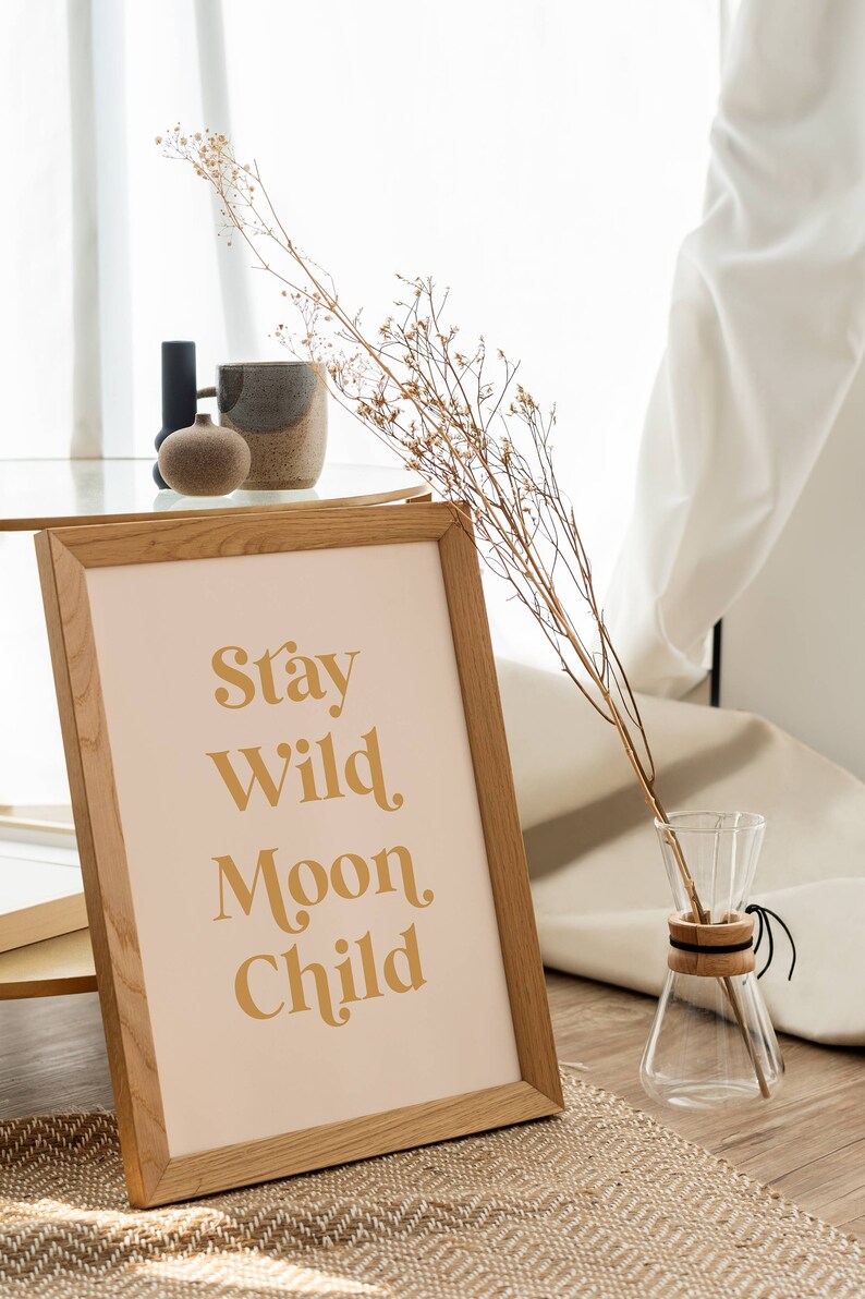 May include: A framed print with the text "Stay Wild Moon Child" in a gold font on a light pink background. The frame is made of light brown wood and is sitting on a woven rug.