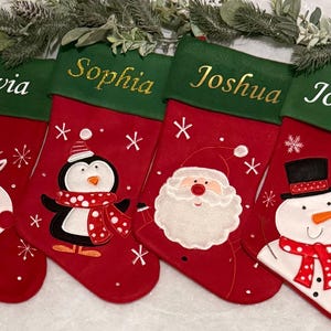 May include: Four red Christmas stockings with green tops and embroidered names: Olivia, Sophia, Joshua, and Joshua. Each stocking features a different embroidered design: a reindeer, a penguin, Santa Claus, and a snowman.