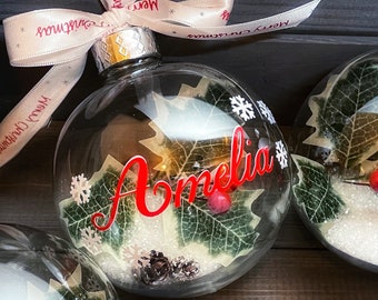 May include: Clear glass Christmas bauble with a red "Amelia" name inside. The bauble is filled with faux snow, green leaves, and pine cones. The bauble is tied with a white ribbon with the text "Merry Christmas" printed on it.