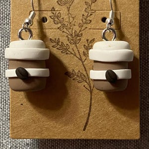 May include: A pair of coffee cup earrings. Each earring has a miniature brown coffee cup with a white rim and a coffee bean accent. The earrings are attached to silver-coloured fish hook ear wires and displayed on a brown card.