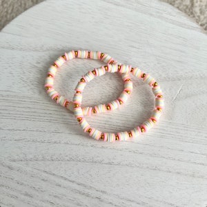 Puede incluir: Dos pulseras hechas de cuentas rosadas, blancas y doradas. Las pulseras están apiladas una encima de la otra.