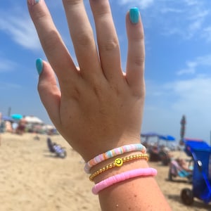 Puede incluir: Una mano con tres pulseras en la muñeca. La pulsera superior es una pulsera de cuentas multicolores. La pulsera del medio es una cadena de oro con un dije de carita sonriente amarilla. La pulsera inferior es una pulsera de cuentas rosadas.