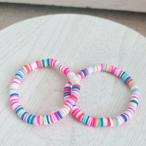 Puede incluir: Dos pulseras coloridas hechas de cuentas redondas y planas apiladas. Las cuentas son en tonos de rosa, azul, blanco y naranja.