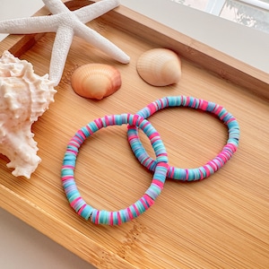 Puede incluir: Dos pulseras coloridas hechas de cuentas redondas de plástico apiladas en tonos de rosa, azul y turquesa. Las pulseras están en una bandeja de madera con conchas marinas y una estrella de mar.